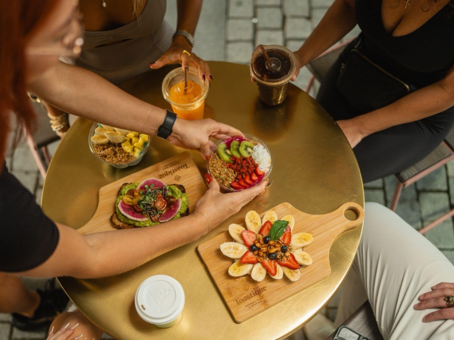Friends sharing a healthy morning routine with avocado toast, açaí bowl, peanut butter banana toast, and fresh juice at Toastique Tampa outdoor table