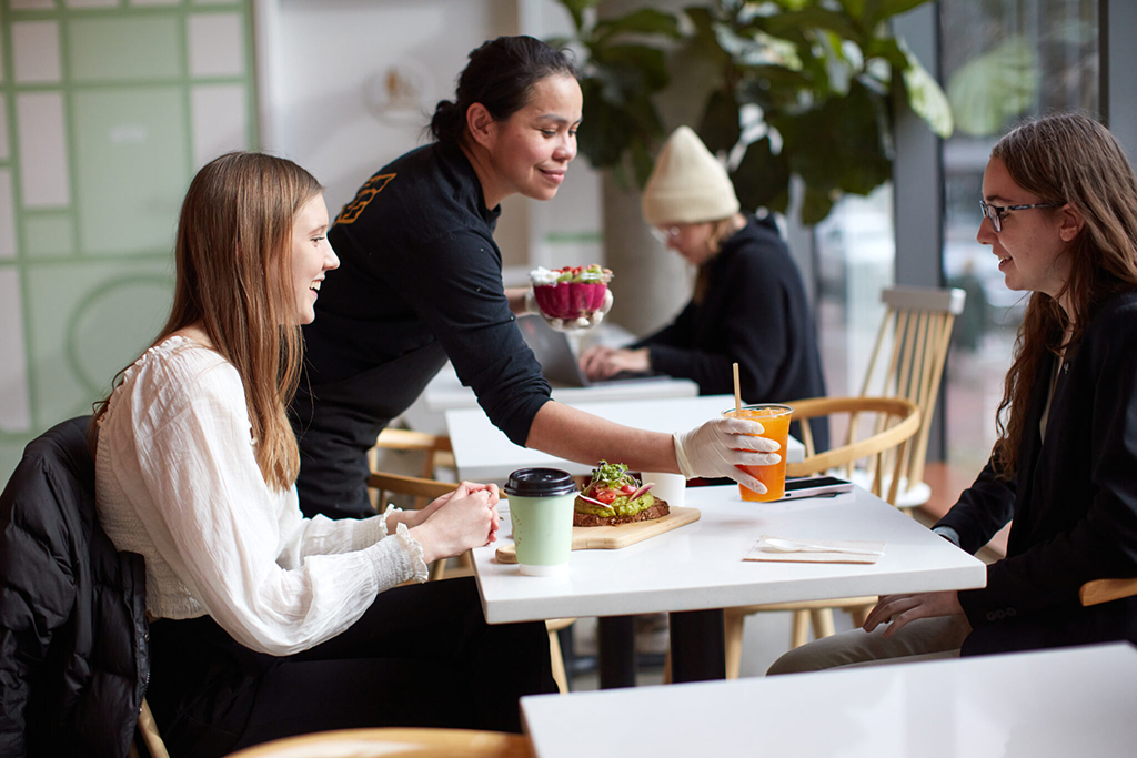 A team member serves vibrant gourmet toasts and fresh juice to smiling guests at a bright Toastique café in Addison, Texas — a modern, welcoming breakfast spot for franchise owners to be proud of.