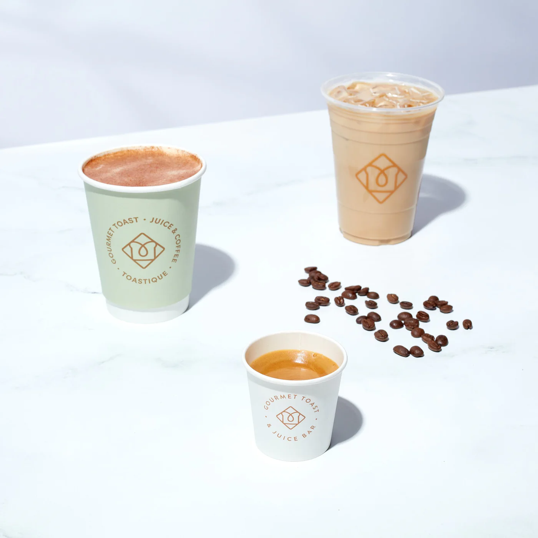 Toastique coffee drinks, including a hot latte, iced coffee, and an espresso shot, on a marble countertop with scattered coffee beans