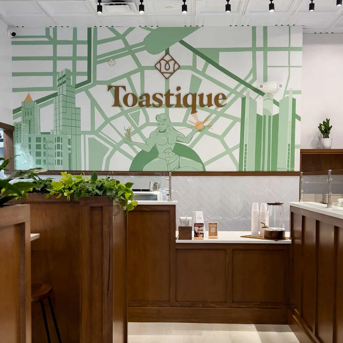 Toastique café interior near Campus Martius in downtown Detroit featuring Spirit of Detroit mural, warm wood accents, and fresh plants at the counter.