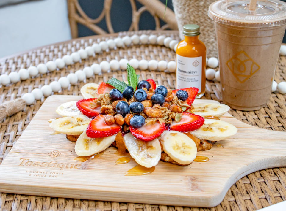 Toastique gourmet toast franchise concept featuring almond butter toast with fresh bananas, strawberries, blueberries, and honey on a signature wooden board with cold-pressed juice.