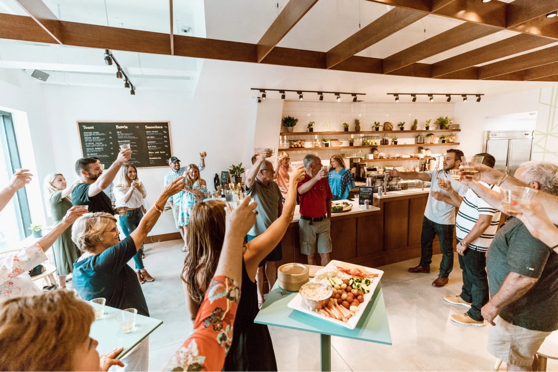 Guests raise glasses and gather around a Toastique café for a lively brunch celebration, with gourmet toast boards, fresh ingredients, and a warm, modern interior in the background.