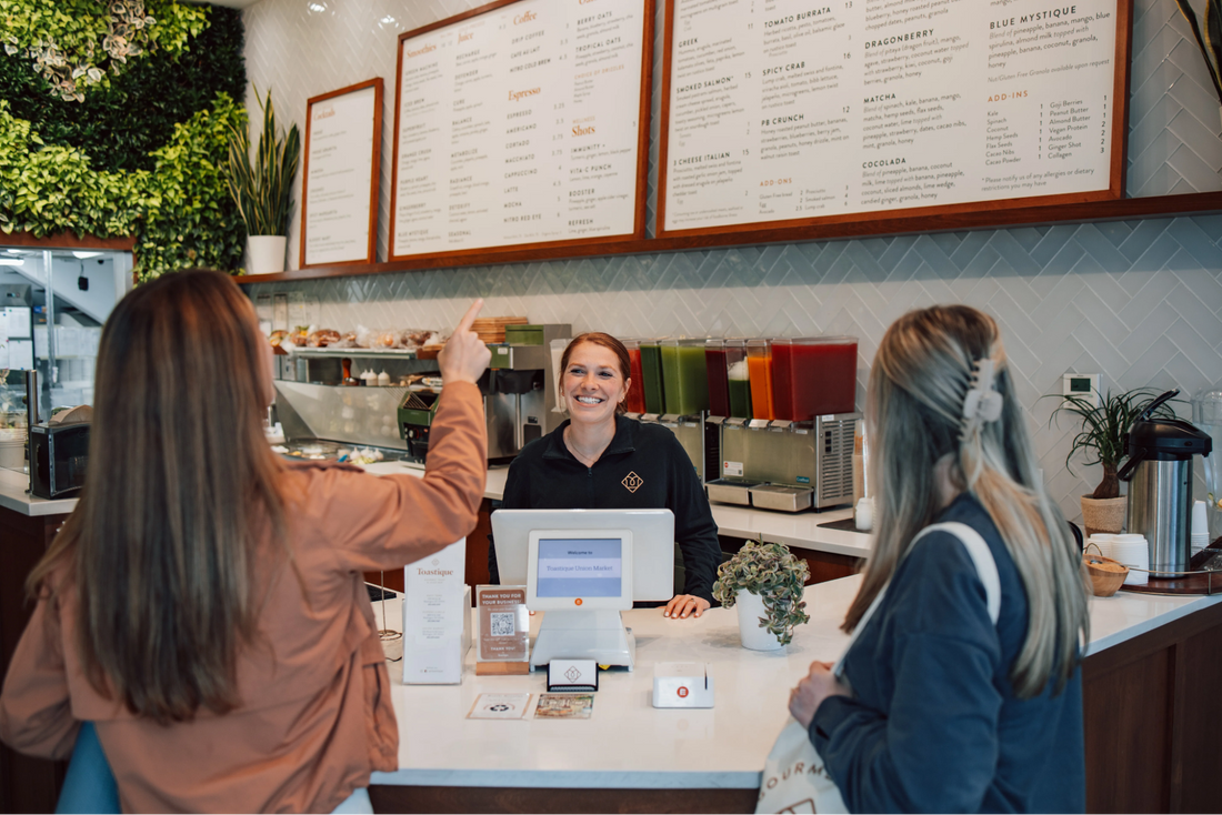 Guests ordering at a Toastique café counter with fresh juices, gourmet toast menu boards, and a smiling team member providing friendly service.