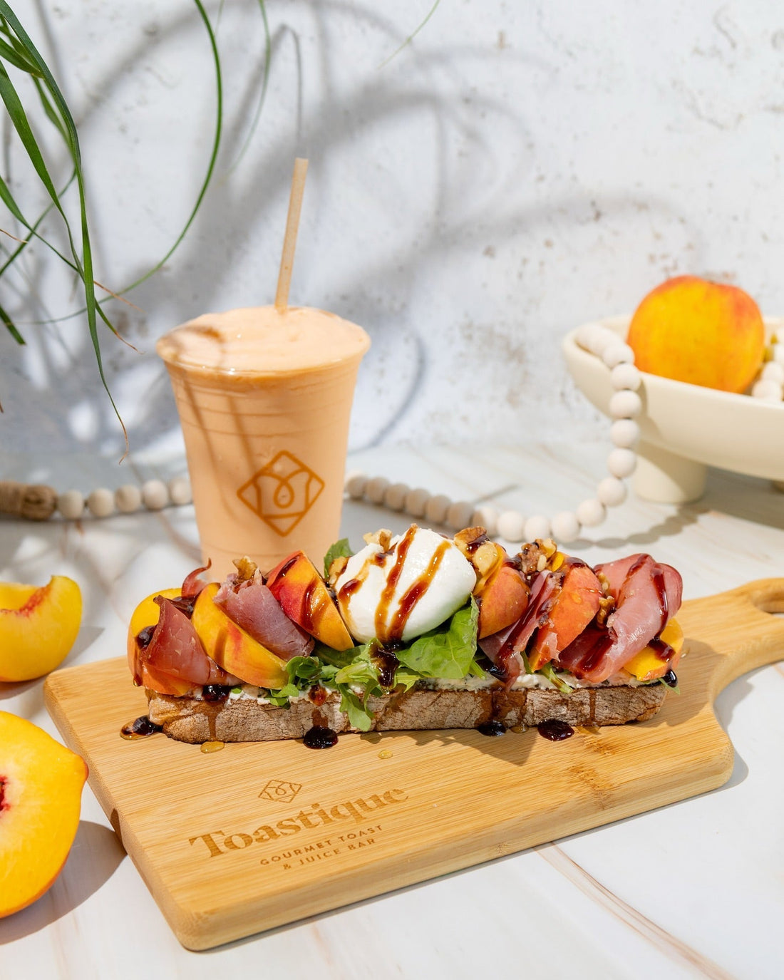 Gourmet peach and prosciutto toast with fresh greens, burrata, walnuts, and balsamic glaze served on rustic bread, paired with a refreshing peach smoothie at Toastique in Arlington, VA.