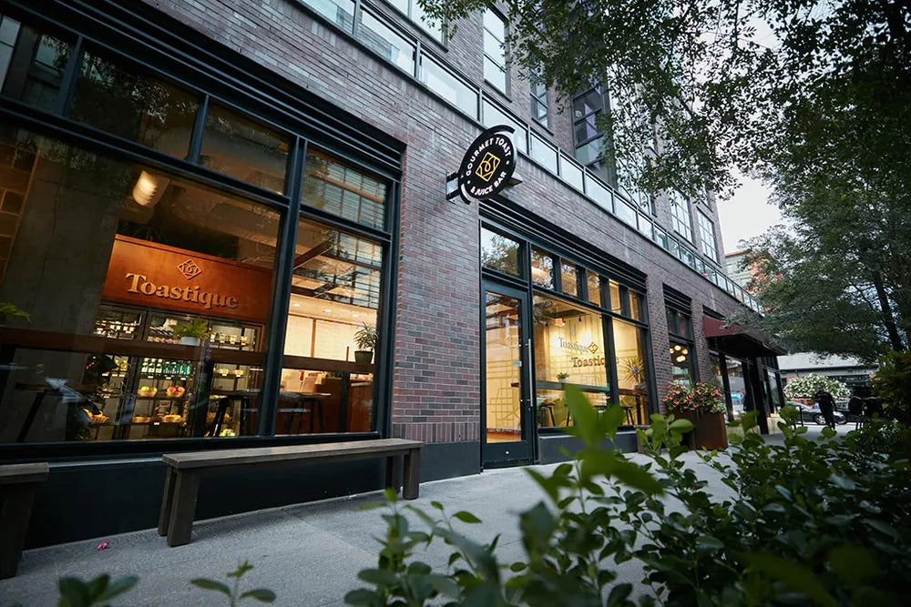 A Toastique cafe at dusk, its lit brand sign and window seating fronting a brick building