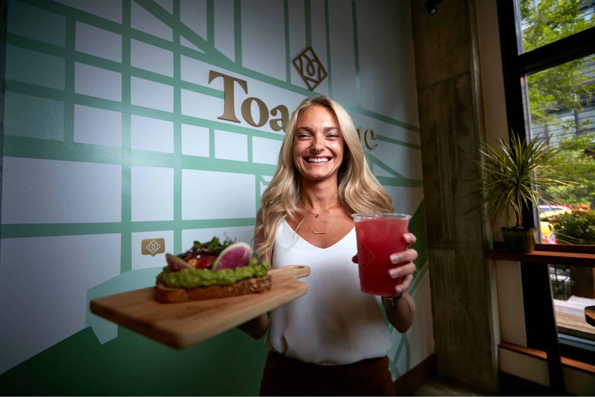 Smiling team member holding gourmet toast and fresh juice inside a Toastique acai bowl franchise café, showcasing vibrant menu and welcoming atmosphere.
