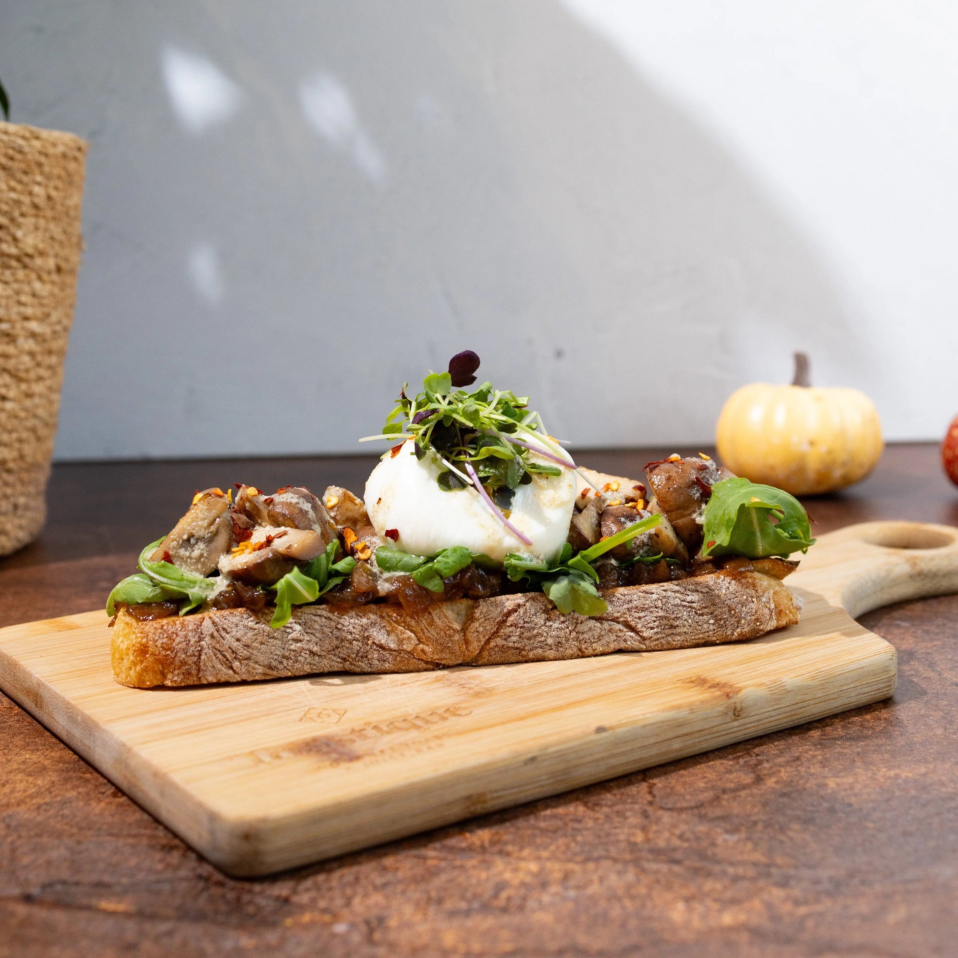 Toasted rustico bread, roasted garlic onion jam, arugula, roasted
mushrooms, burrata, caesar dressing, chili flakes, maldon salt,
topped with microgreens , served on a wooden cutting board on a rustic wooden table with a small pumpkin and plant in the background.