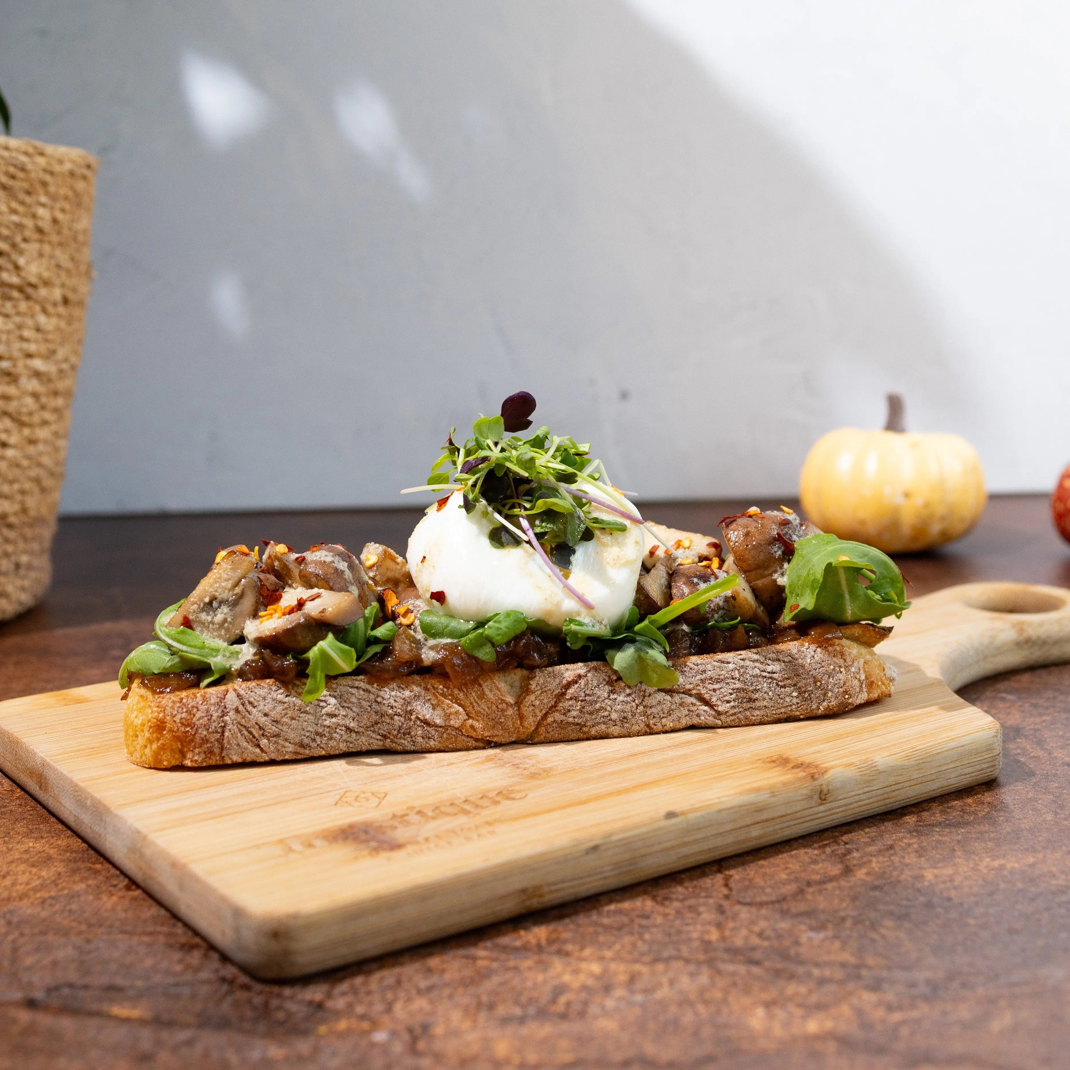 Toasted rustico bread, roasted garlic onion jam, arugula, roasted
mushrooms, burrata, caesar dressing, chili flakes, maldon salt,
topped with microgreens , served on a wooden cutting board on a rustic wooden table with a small pumpkin and plant in the background.