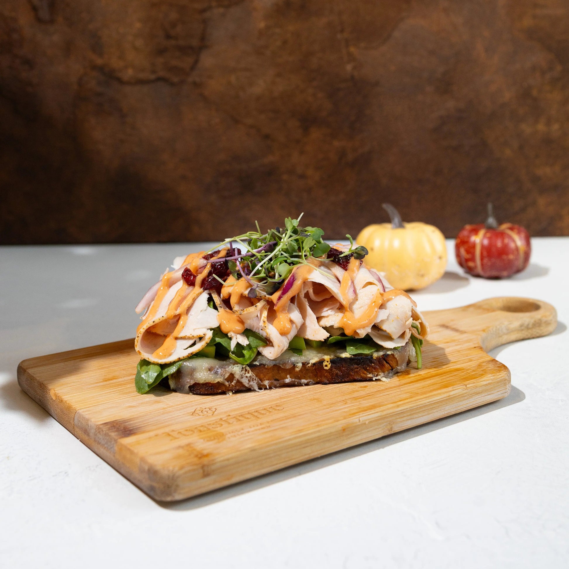 oasted walnut raisin bread, chipotle aioli drizzle, melted swiss
and fontina, granny smith apples, arugula, sliced turkey, red
onions, dried cranberries, topped with microgreens, served on a wooden cutting board on a rustic wooden table with a small pumpkin in the background.