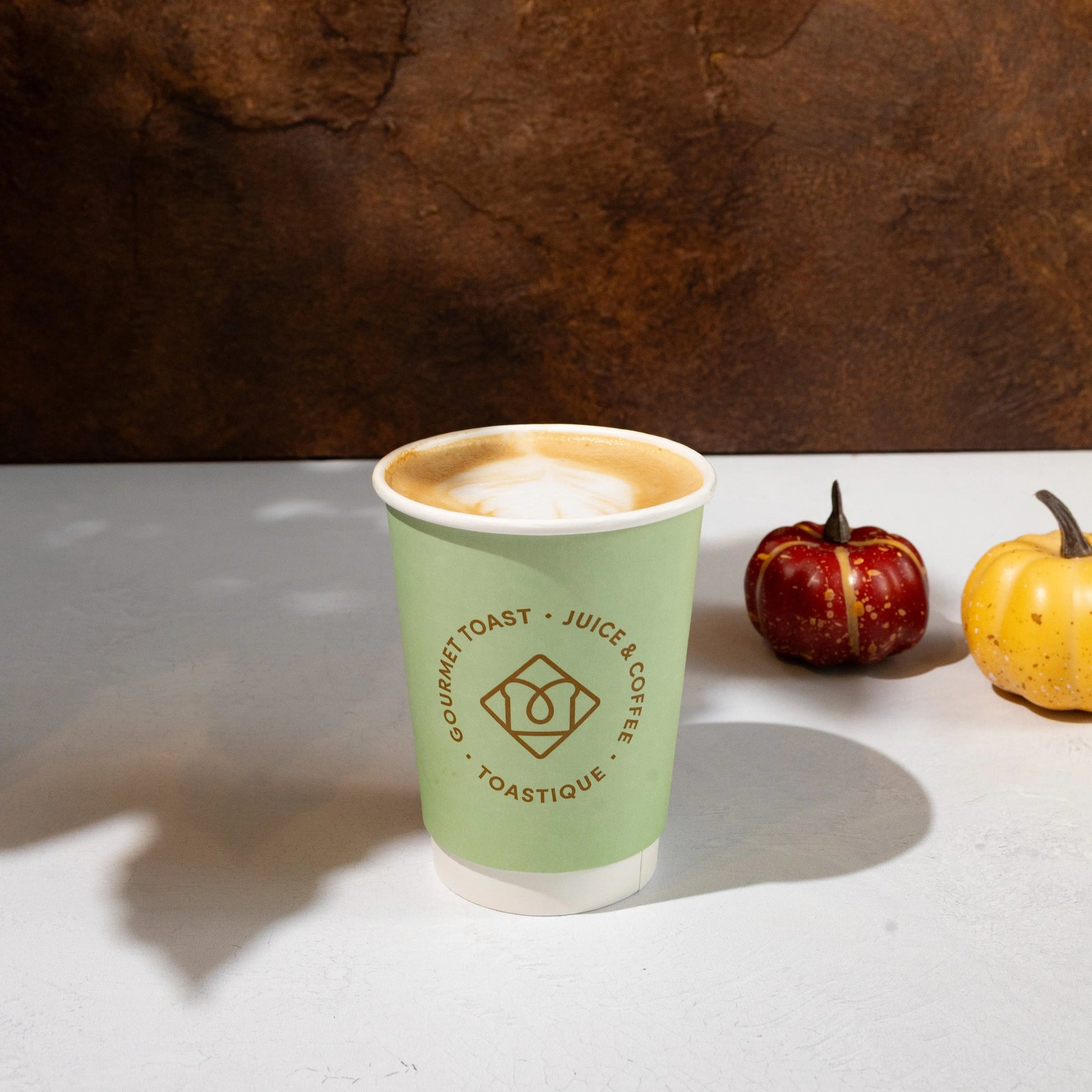 Toastique Pumpkin Spice Latte in a green branded cup, pictured with mini pumpkins on a white table against a warm brown backdrop.