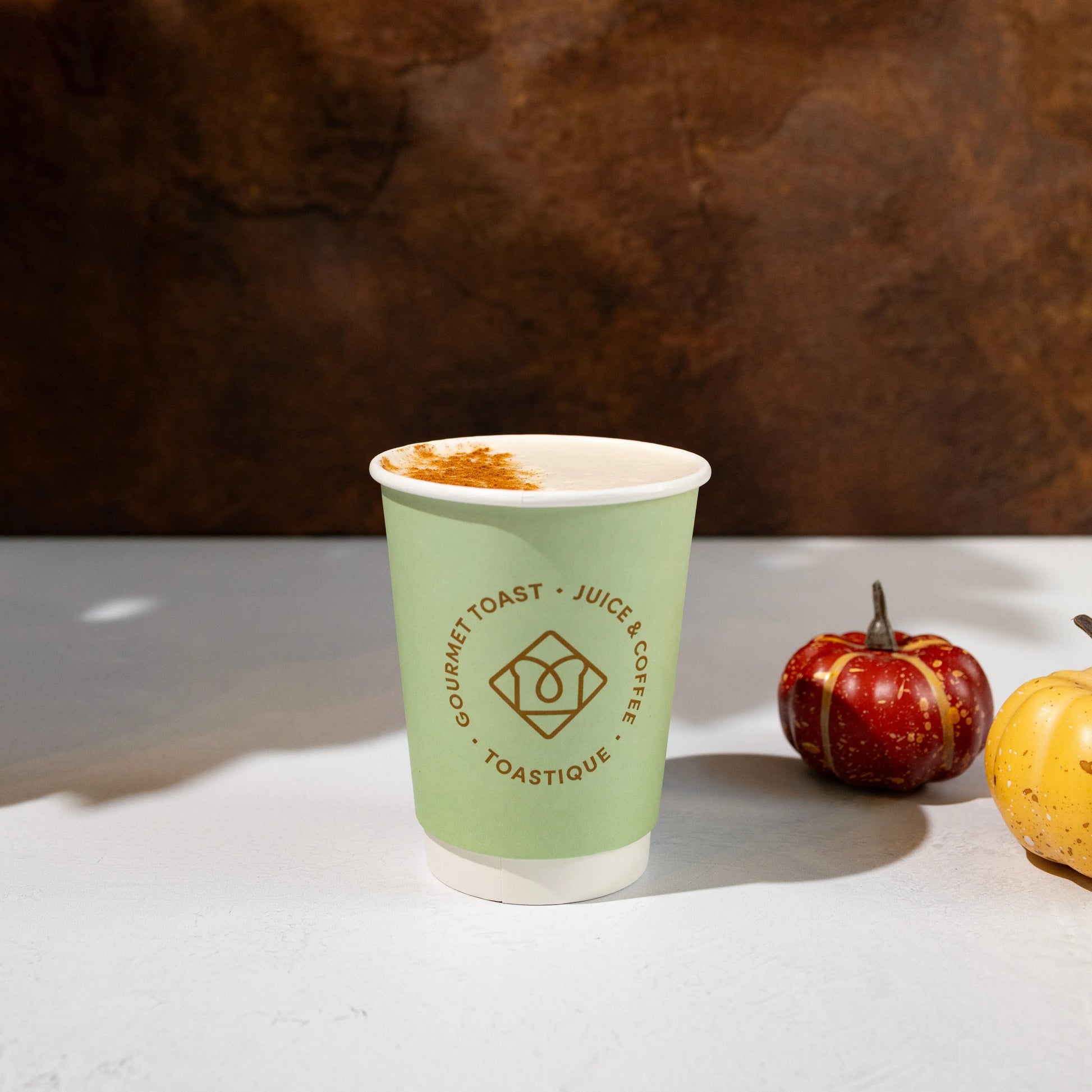 Toastique Caramel Apple Chai Latte in a green branded cup, topped with cinnamon and pictured with mini pumpkins on a white table against a warm brown backdrop.
