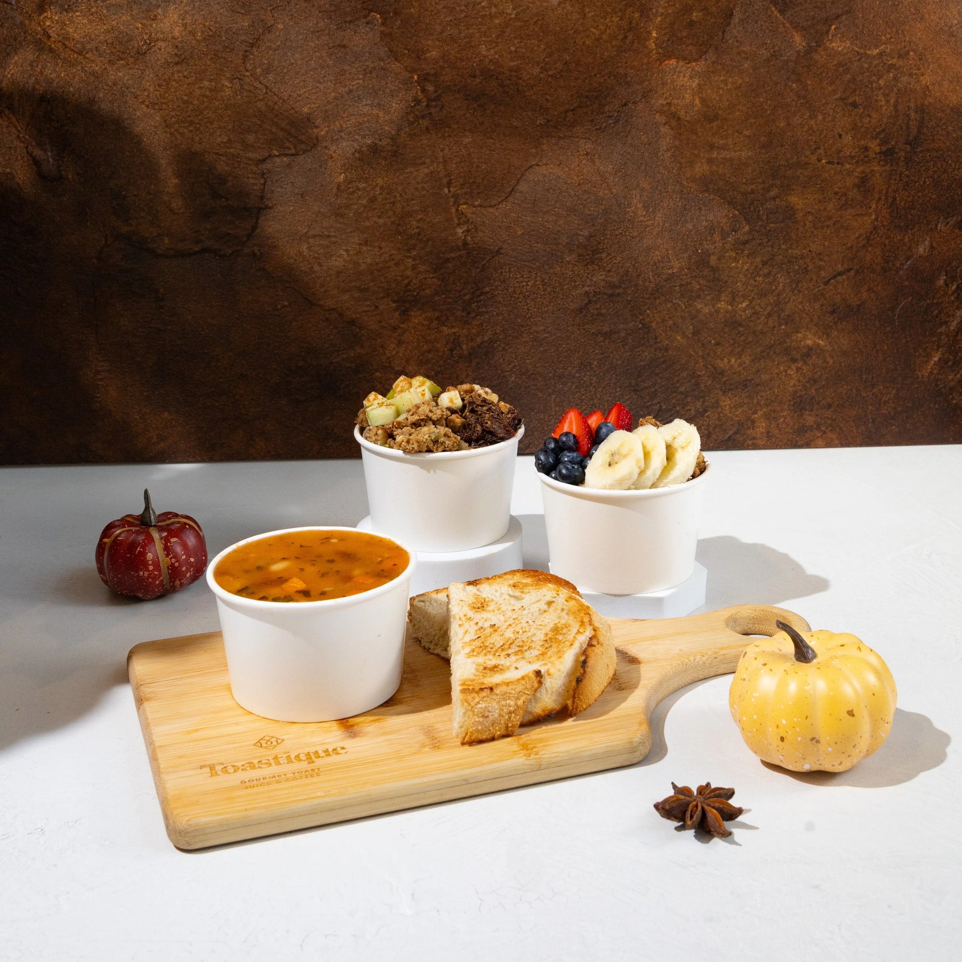 Toastique Hot Berry Oats, Apple Spice Oats and cup of soup on a Toastique wooden board with pumpkins in the background.