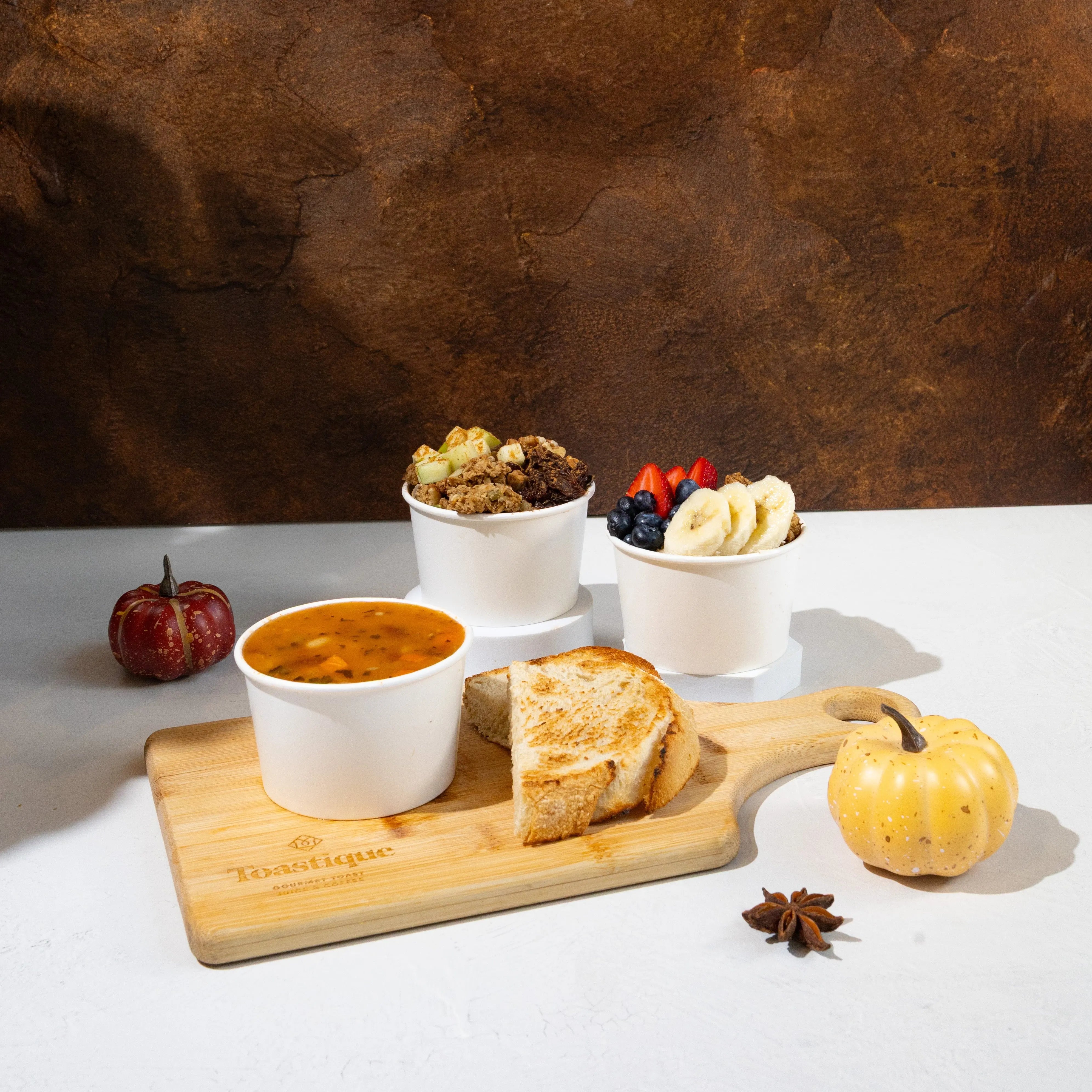 Toastique Hot Berry Oats, Apple Spice Oats and cup of soup on a Toastique wooden board with pumpkins in the background.